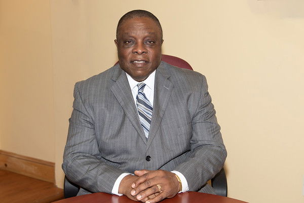 A Black man in a gray pinstriped suit and striped tie sits at a wooden table with his hands clasped in front of him. Stanley King looks directly at the camera with a serious expression against a beige wall background.